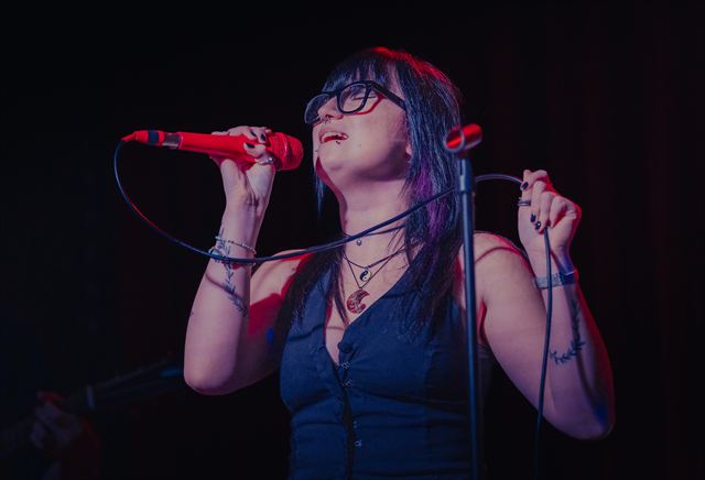 A singer with glasses and a red microphone, eyes closed mid-note under blue light