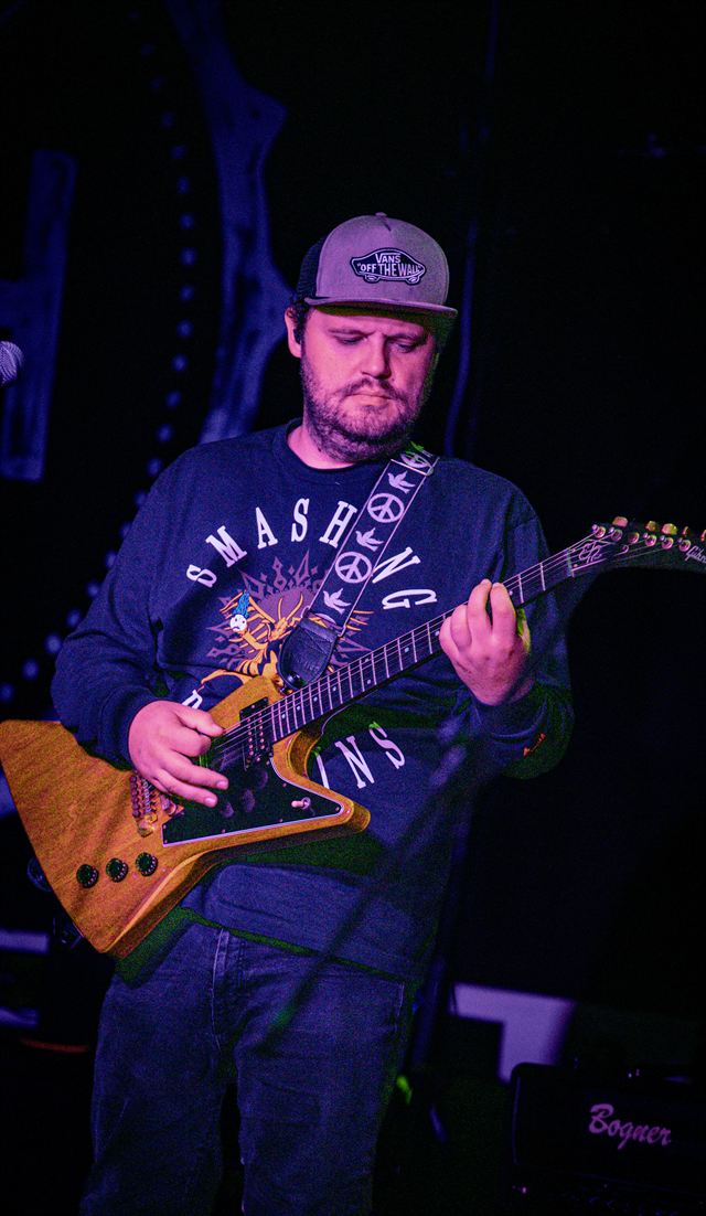 A guitarist in a cap and hoodie playing an angular electric guitar