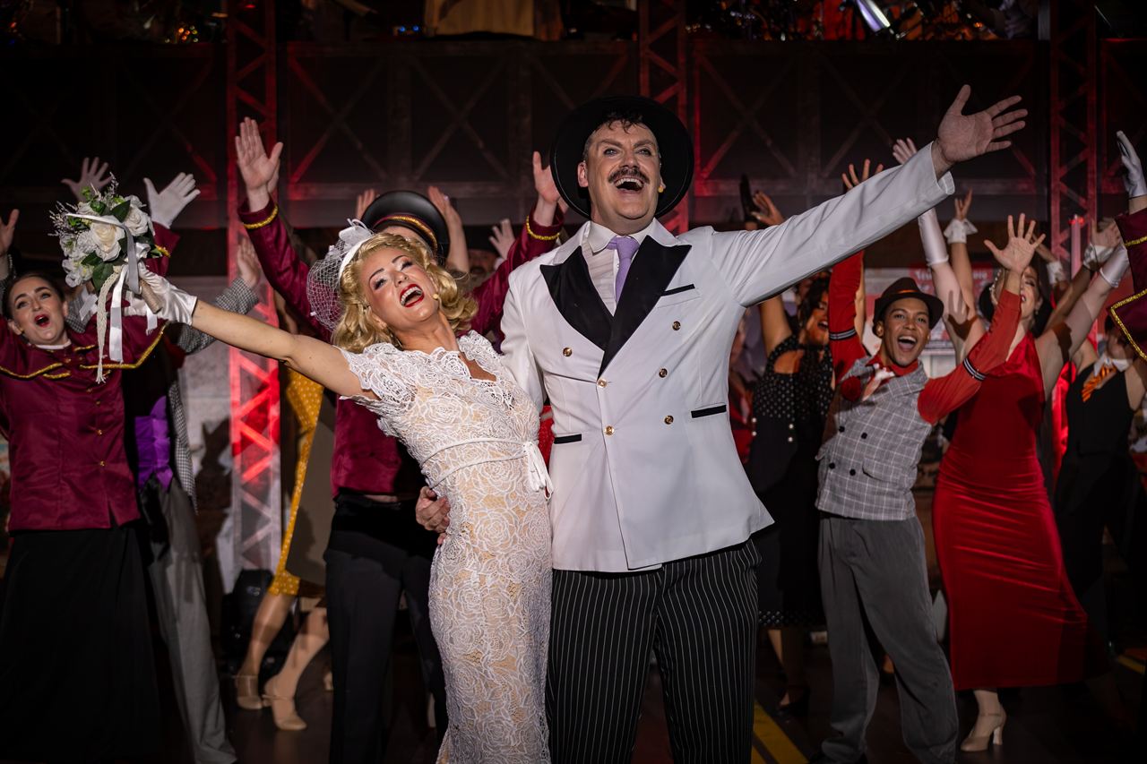 The full cast throwing their arms up in the finale, led by a man in a white suit