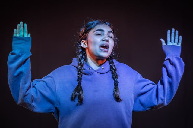 A performer with long braids in a lilac hoodie singing with her eyes closed and both hands raised under blue stage light