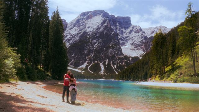Two people on the shore of a turquoise mountain lake surrounded by pine forest in the Dolomites
