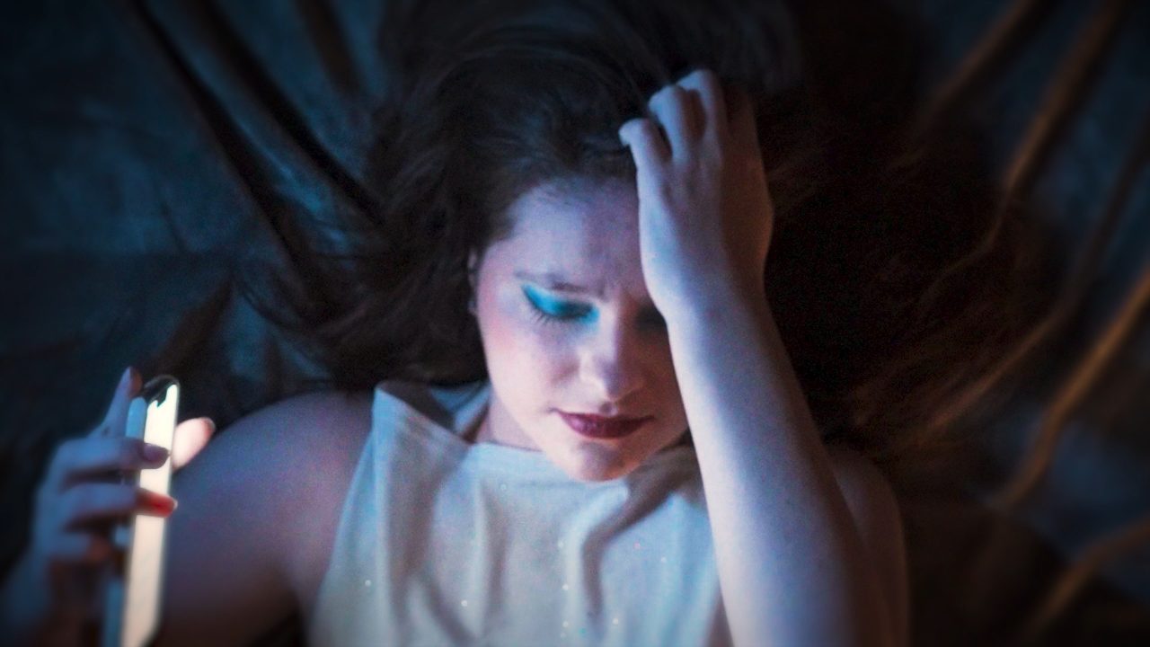 A woman lying in bed lit by cold blue light, holding her phone